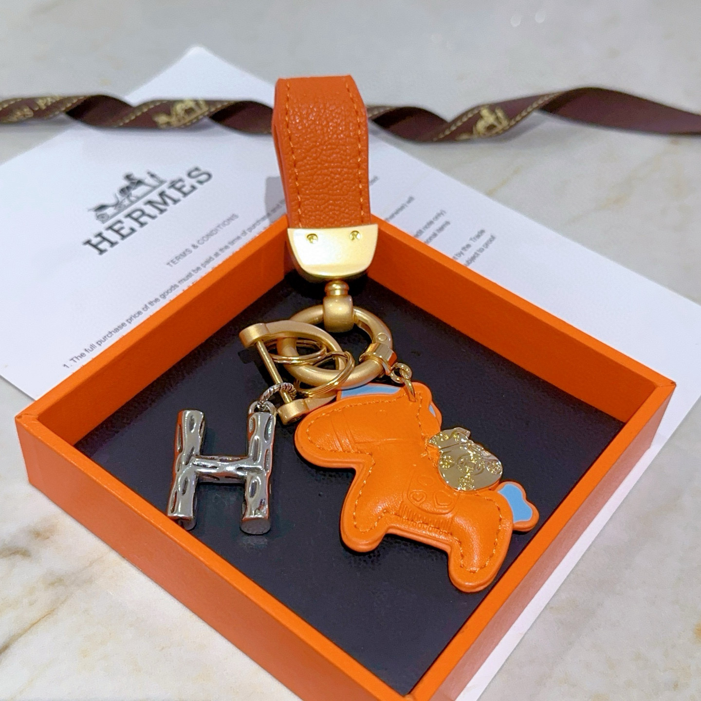 Colgante Hermes Rodeo Pegase H Logo Key Charm Cuero naranja con detalles en azul claro Herrajes metálicos en tono dorado y plateado Longitud total de 8 cm