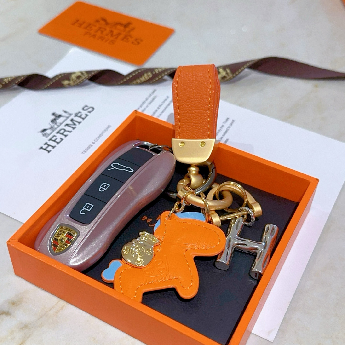 Colgante Hermes Rodeo Pegase H Logo Key Charm Cuero naranja con detalles en azul claro Herrajes metálicos en tono dorado y plateado Longitud total de 8 cm