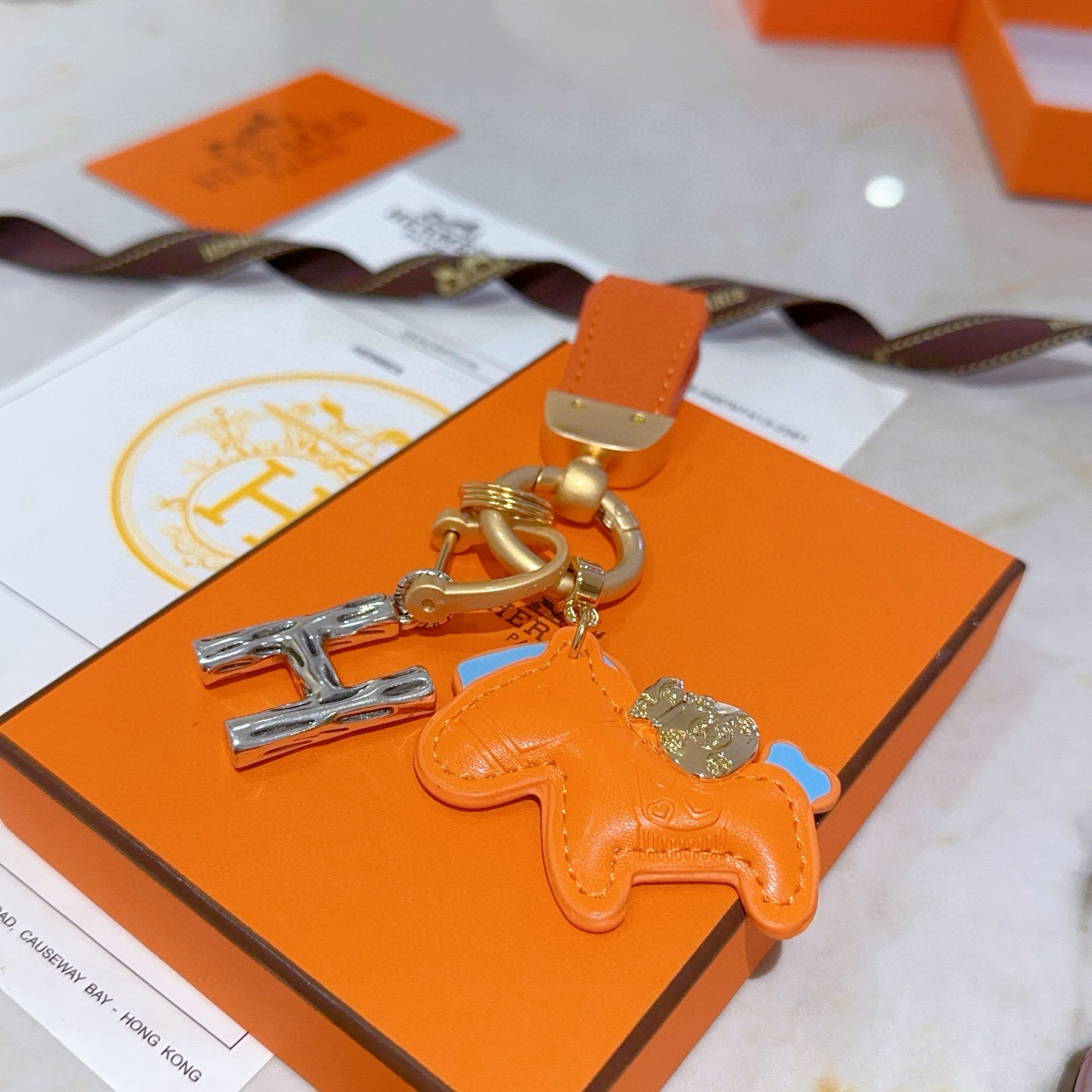 Colgante Hermes Rodeo Pegase H Logo Key Charm Cuero naranja con detalles en azul claro Herrajes metálicos en tono dorado y plateado Longitud total de 8 cm