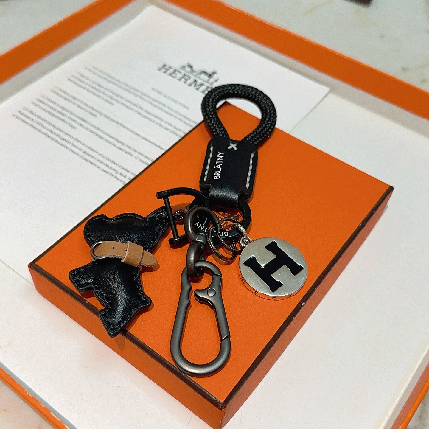 Hermes H Logo Bulldog Charm Chaveiro pingente Cordão trançado preto Couro preto com detalhes em couro bronzeado Hardware em metal prateado com esmalte preto Logotipo H 12 cm de comprimento total