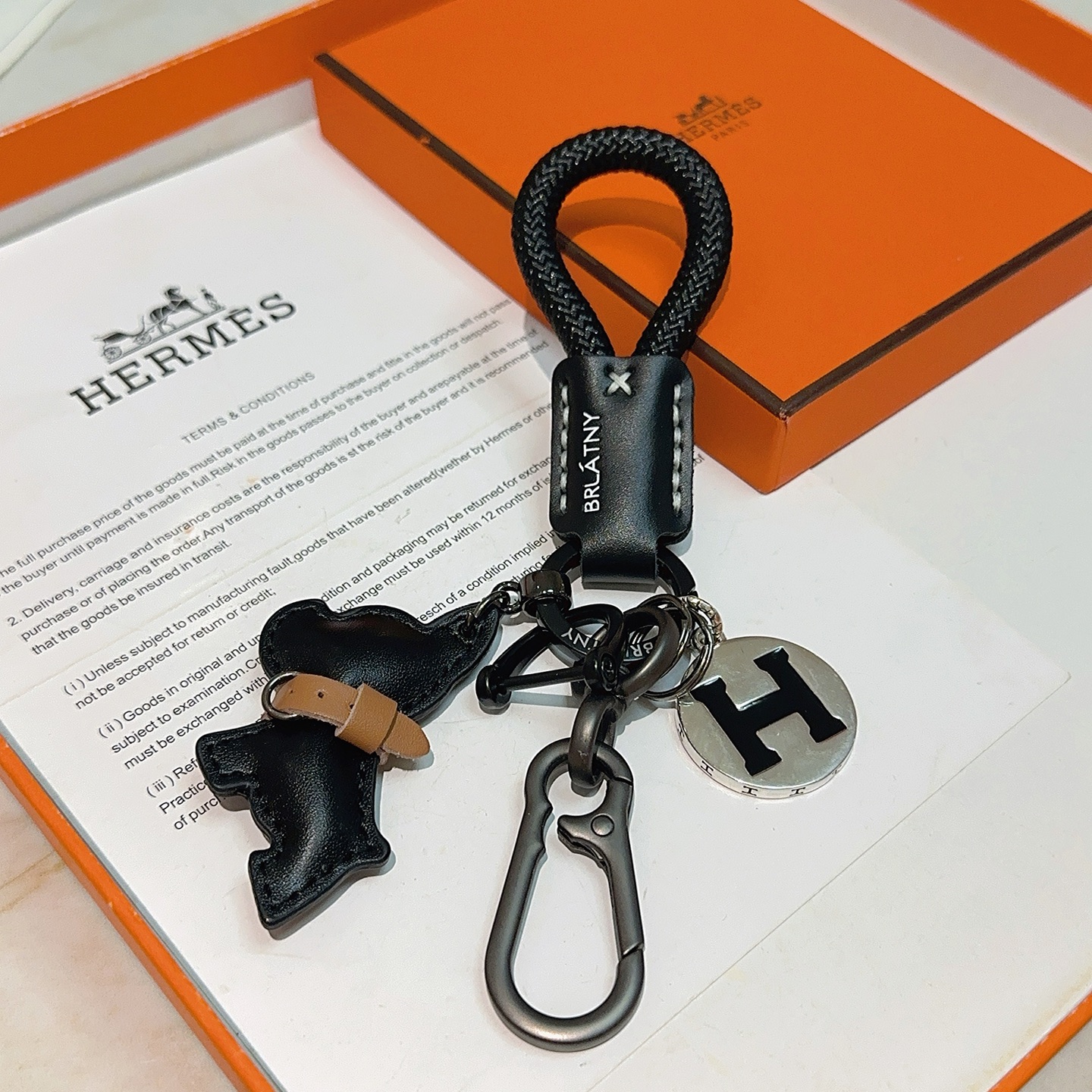 Hermes H Logo Bulldog Charm Chaveiro pingente Cordão trançado preto Couro preto com detalhes em couro bronzeado Hardware em metal prateado com esmalte preto Logotipo H 12 cm de comprimento total