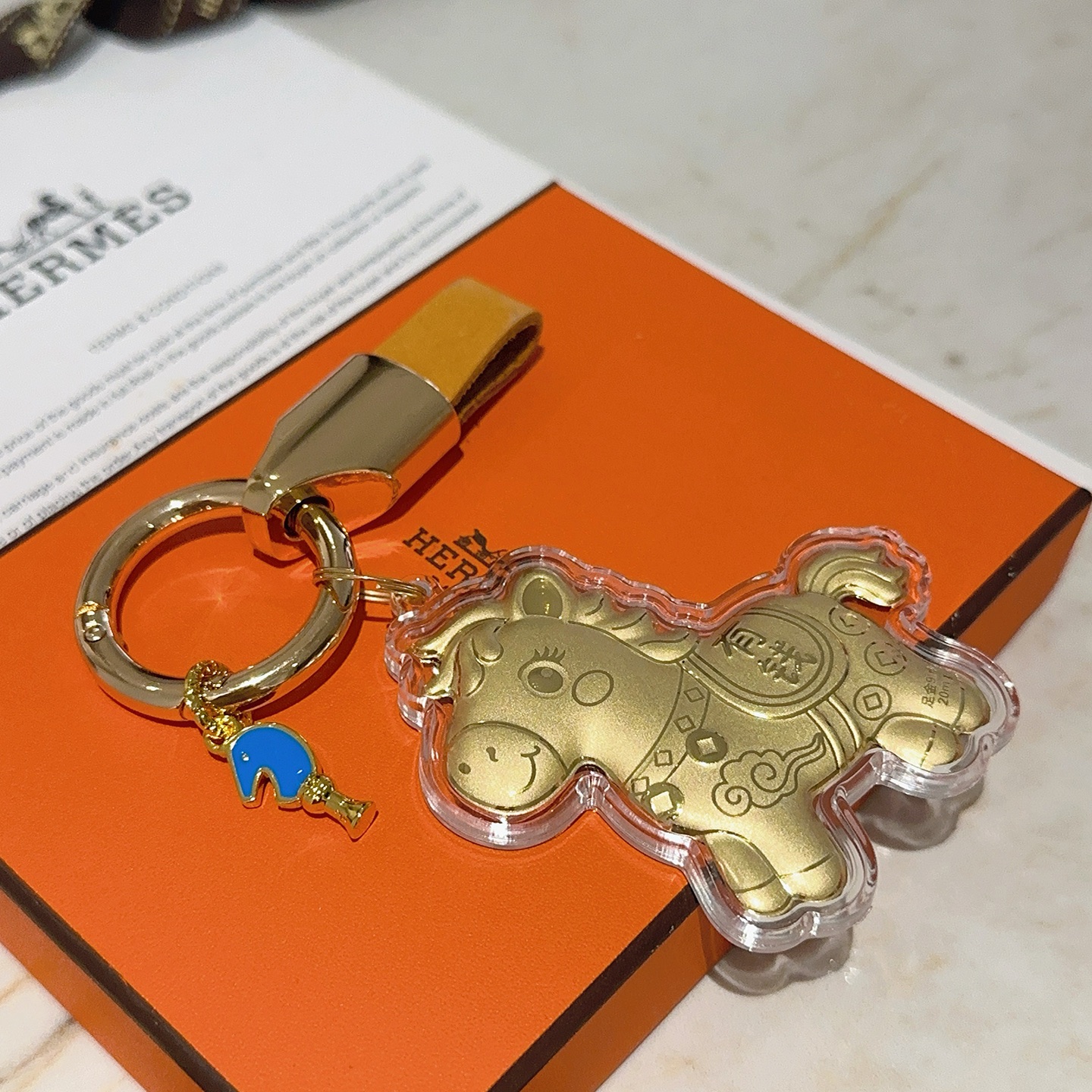 Ciondolo portachiavi Hermes Lucky Horse Charm Metallo color oro con cinturino in pelle arancione accento smaltato blu involucro protettivo trasparente sul ciondolo cavallo dorato Lunghezza totale 9 cm