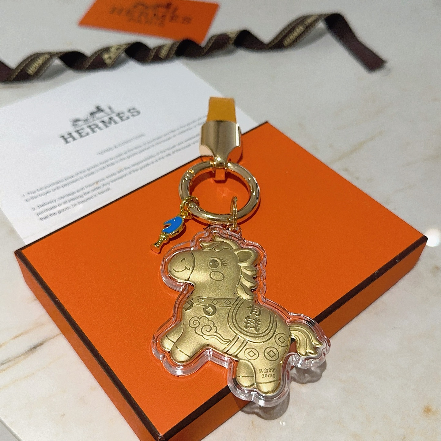 Ciondolo portachiavi Hermes Lucky Horse Charm Metallo color oro con cinturino in pelle arancione accento smaltato blu involucro protettivo trasparente sul ciondolo cavallo dorato Lunghezza totale 9 cm