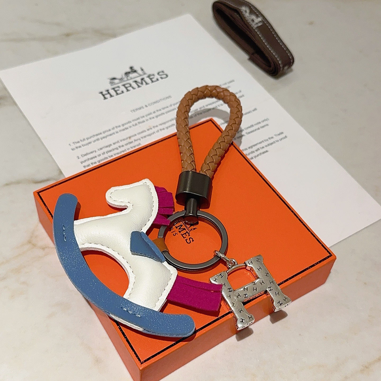 Ciondolo chiave Hermes con cavallo a dondolo H Logo Cinturino in pelle intrecciata marrone Hardware in canna di fucile Ciondolo H color argento Cavallo a dondolo in pelle bianca con sella a bilanciere blu accenti di coda con criniera in pelle fucsia Lunghezza totale 10 cm
