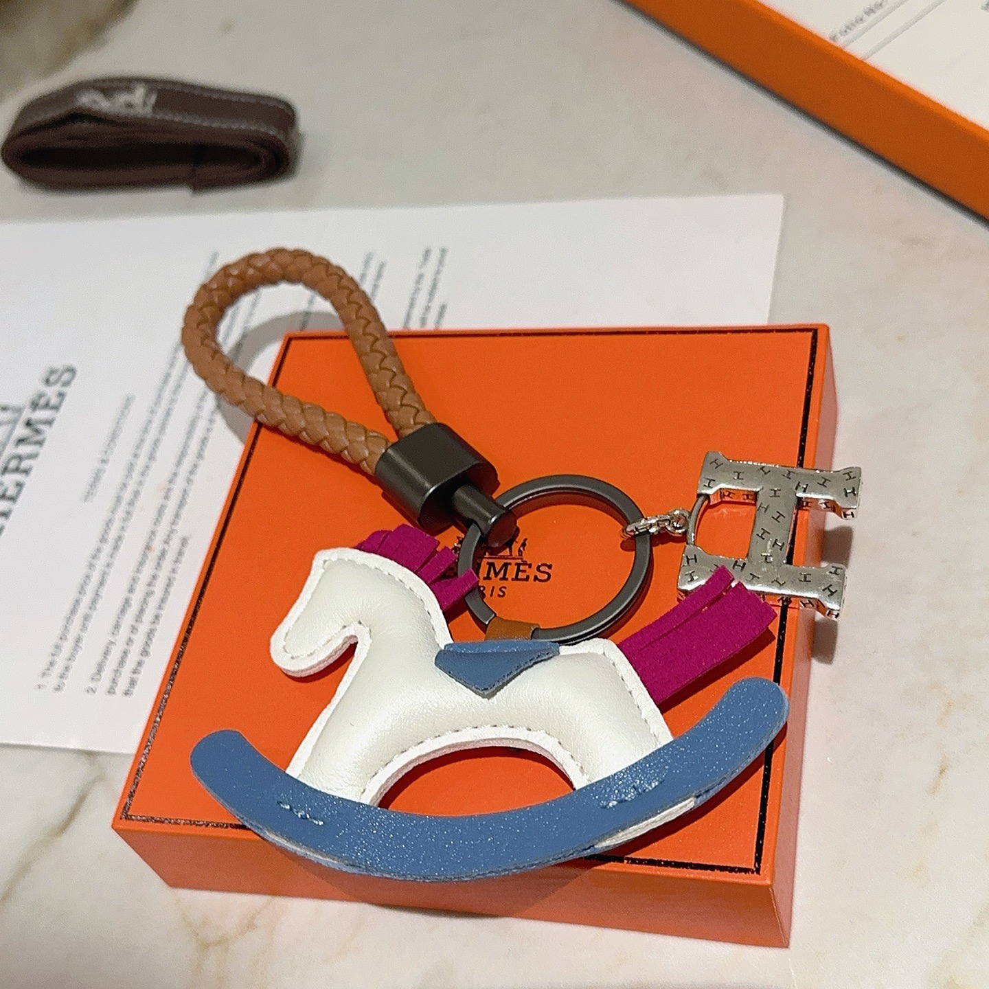 Ciondolo chiave Hermes con cavallo a dondolo H Logo Cinturino in pelle intrecciata marrone Hardware in canna di fucile Ciondolo H color argento Cavallo a dondolo in pelle bianca con sella a bilanciere blu accenti di coda con criniera in pelle fucsia Lunghezza totale 10 cm