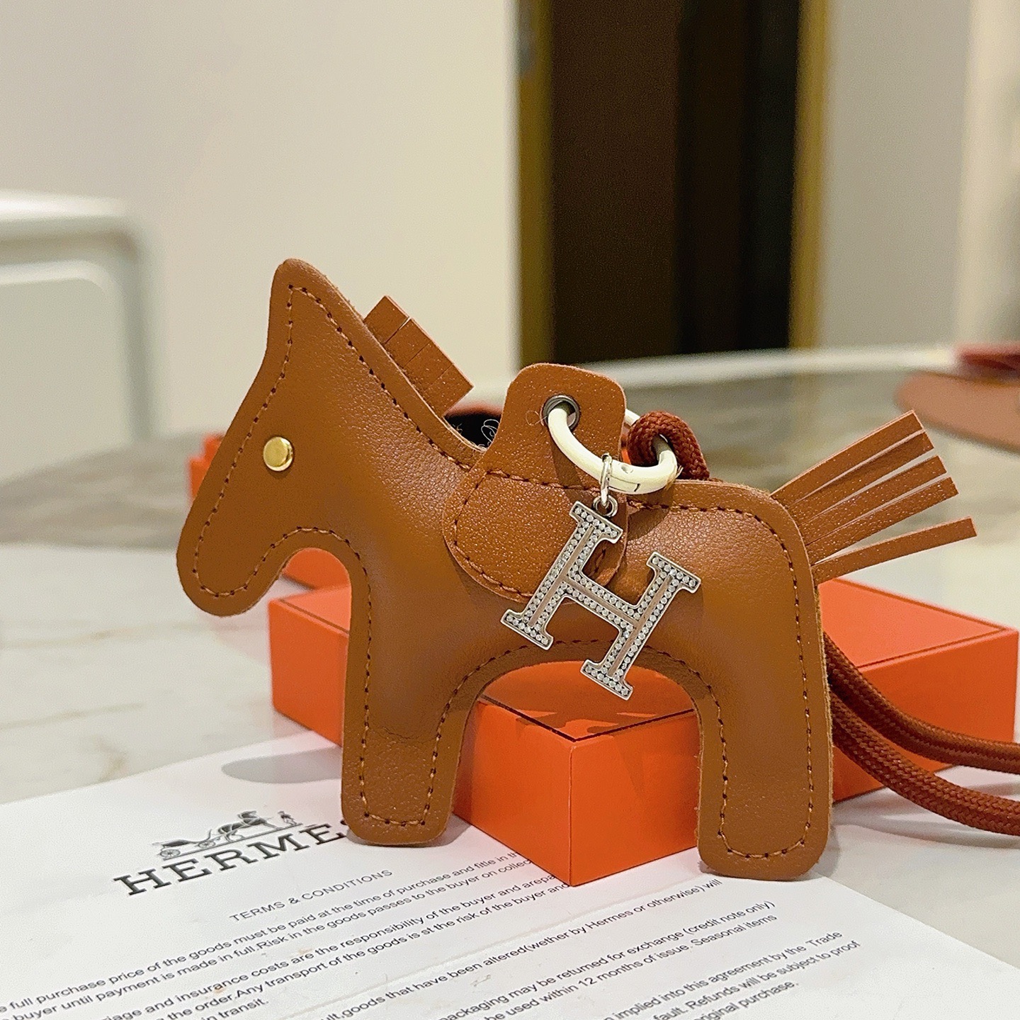 Ciondolo per borsa Hermes Rodeo Pegase Horse con pendente in cristallo H. Pelle marrone oro, metallo color argento con pavé di cristalli trasparenti, hardware bianco sporco, cordone marrone terracotta, lunghezza totale del ciondolo: 8 cm.