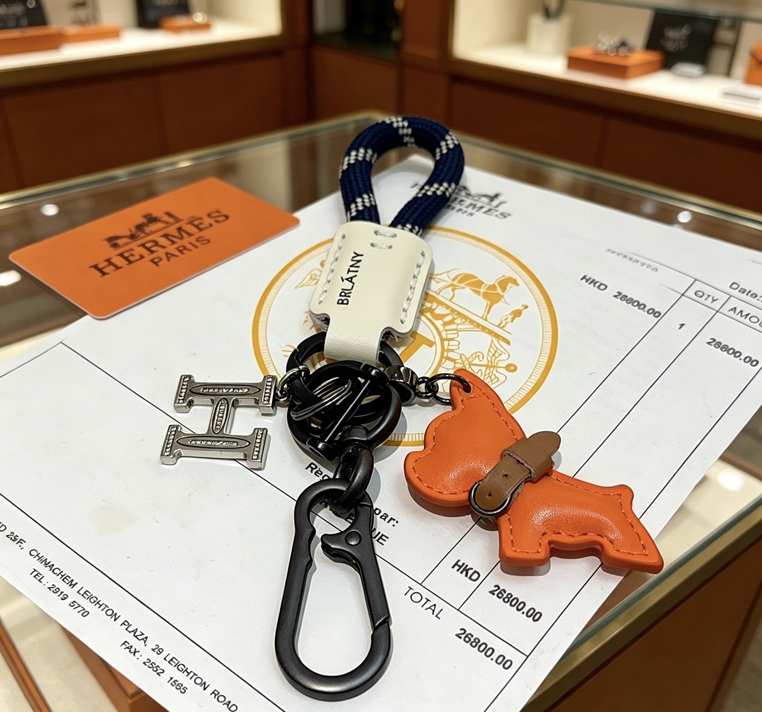 Hermes H Logo Puppy Charm Llavero colgante Herrajes de bronce de cañón Charm de cachorro de cuero naranja Etiqueta de cuero crema Correa tejida azul marino con patrón de cuadros blancos Detalle de collar de cuero marrón 12 cm de longitud total
