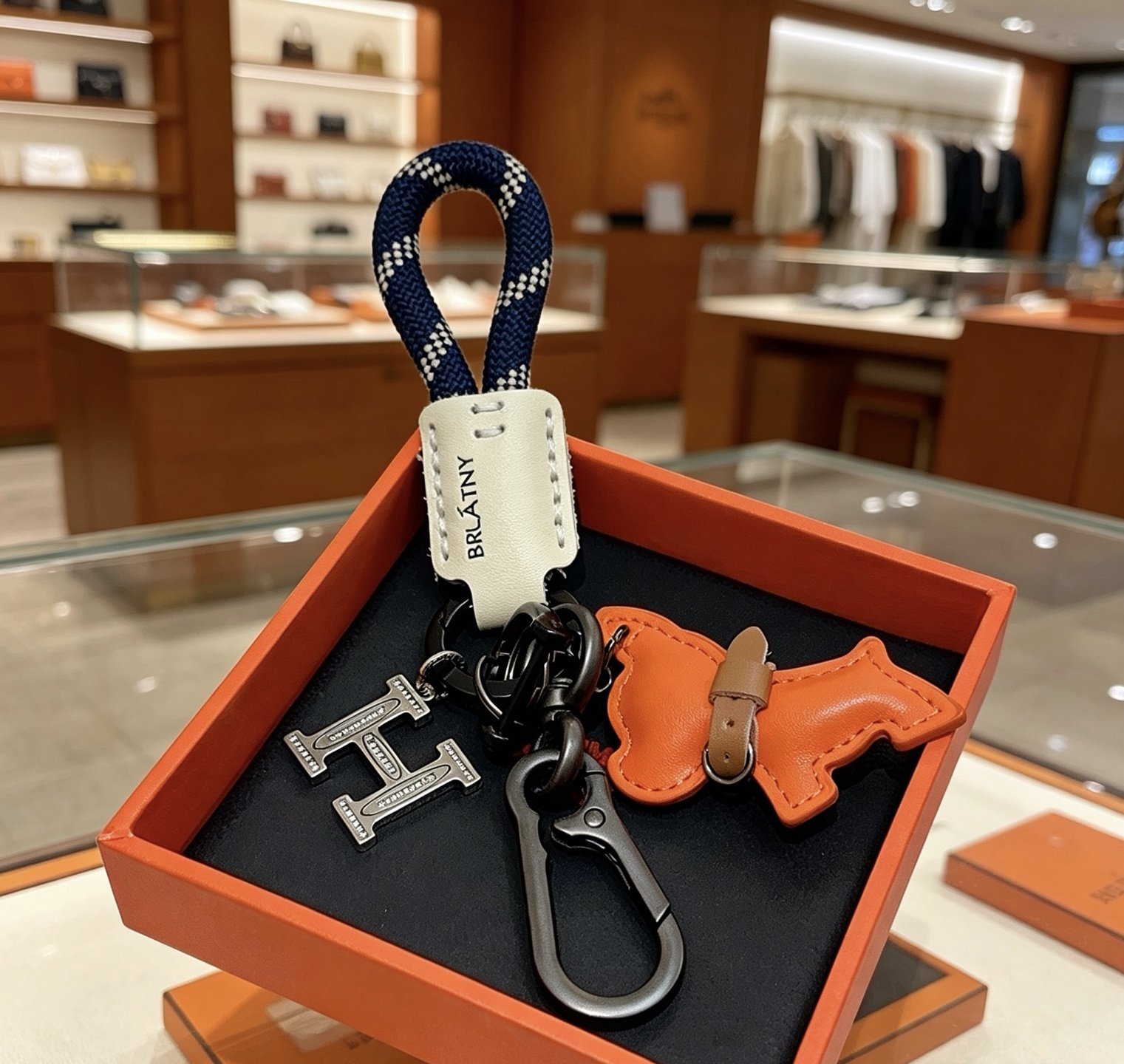 Hermes H Logo Puppy Charm Llavero colgante Herrajes de bronce de cañón Charm de cachorro de cuero naranja Etiqueta de cuero crema Correa tejida azul marino con patrón de cuadros blancos Detalle de collar de cuero marrón 12 cm de longitud total