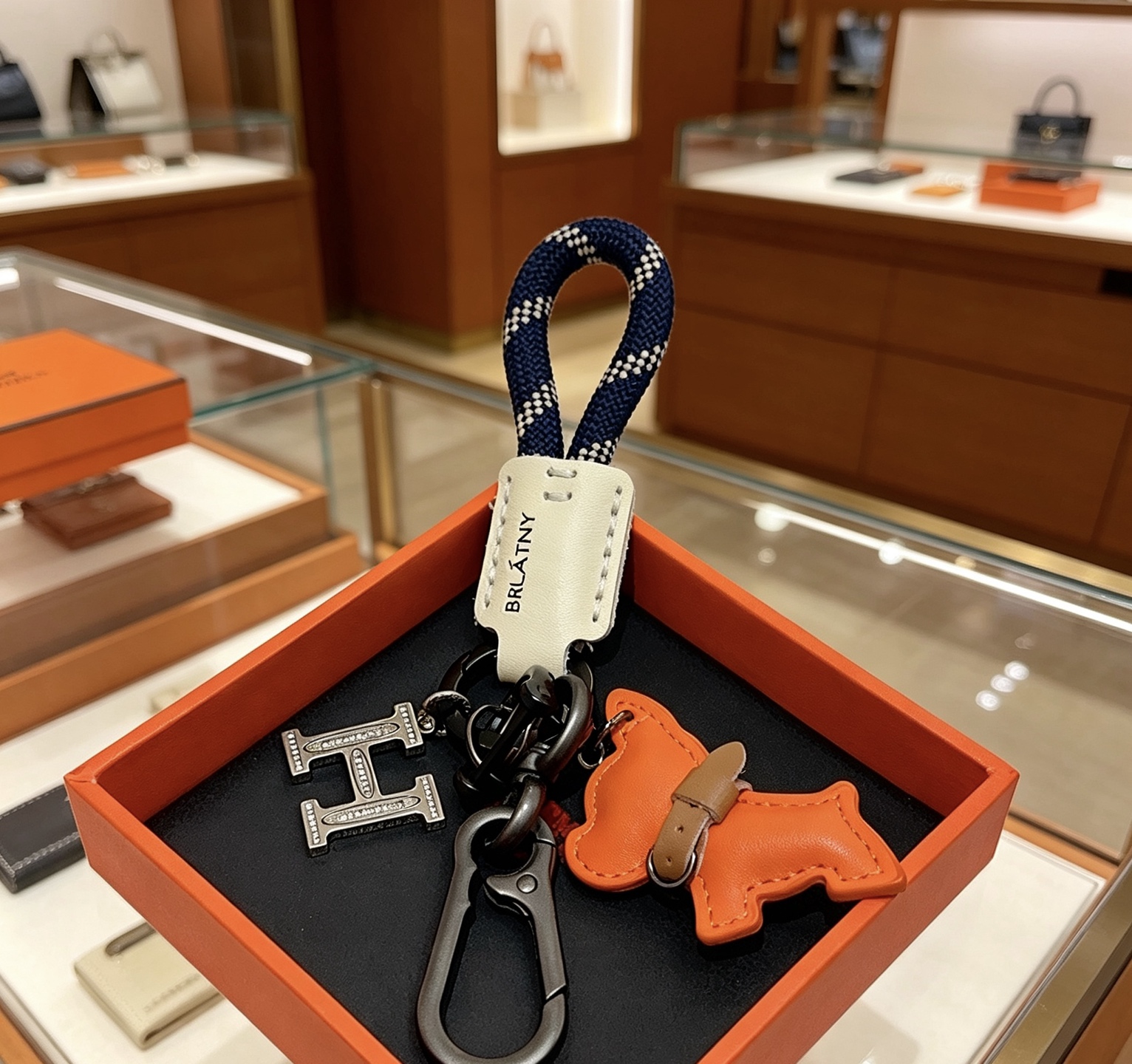 Hermes H Logo Puppy Charm Llavero colgante Herrajes de bronce de cañón Charm de cachorro de cuero naranja Etiqueta de cuero crema Correa tejida azul marino con patrón de cuadros blancos Detalle de collar de cuero marrón 12 cm de longitud total