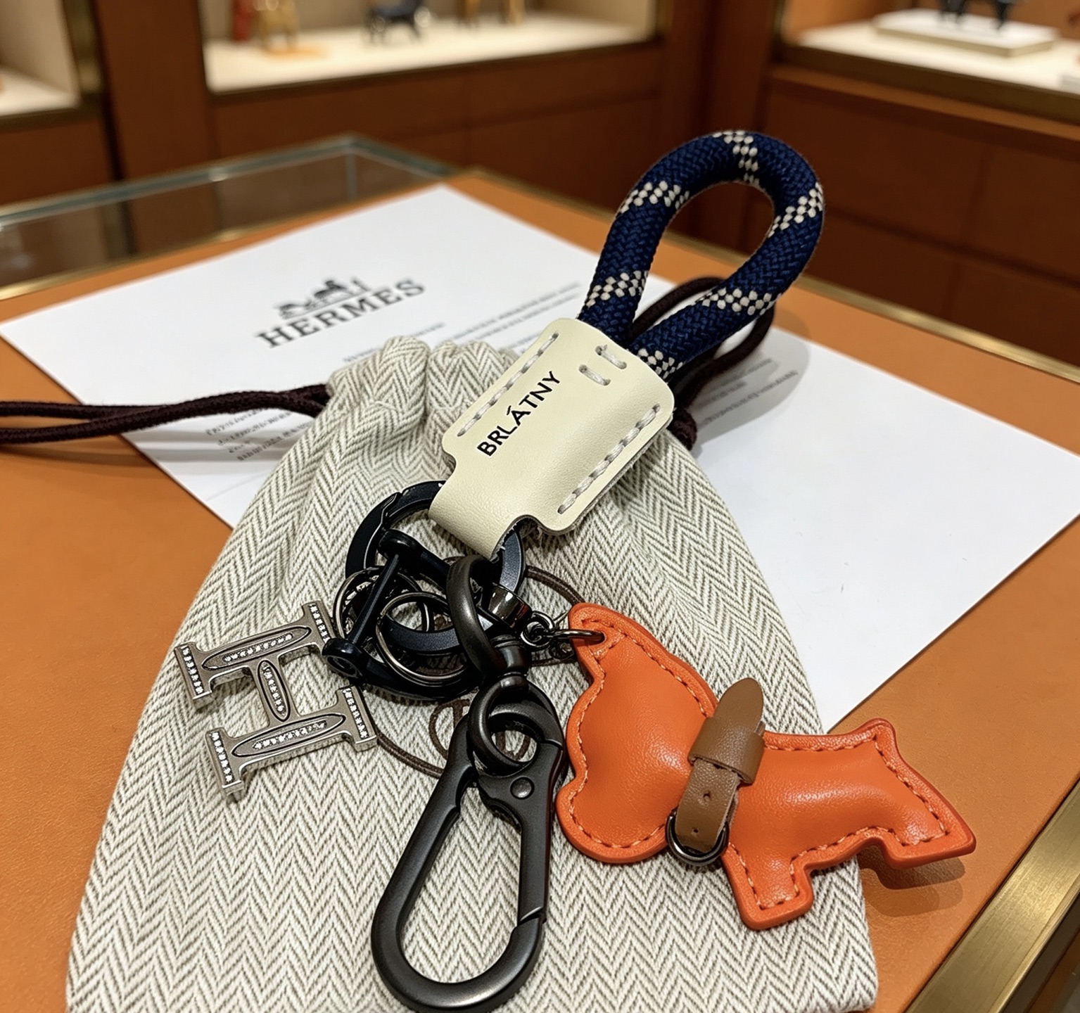 Hermes H Logo Puppy Charm Llavero colgante Herrajes de bronce de cañón Charm de cachorro de cuero naranja Etiqueta de cuero crema Correa tejida azul marino con patrón de cuadros blancos Detalle de collar de cuero marrón 12 cm de longitud total
