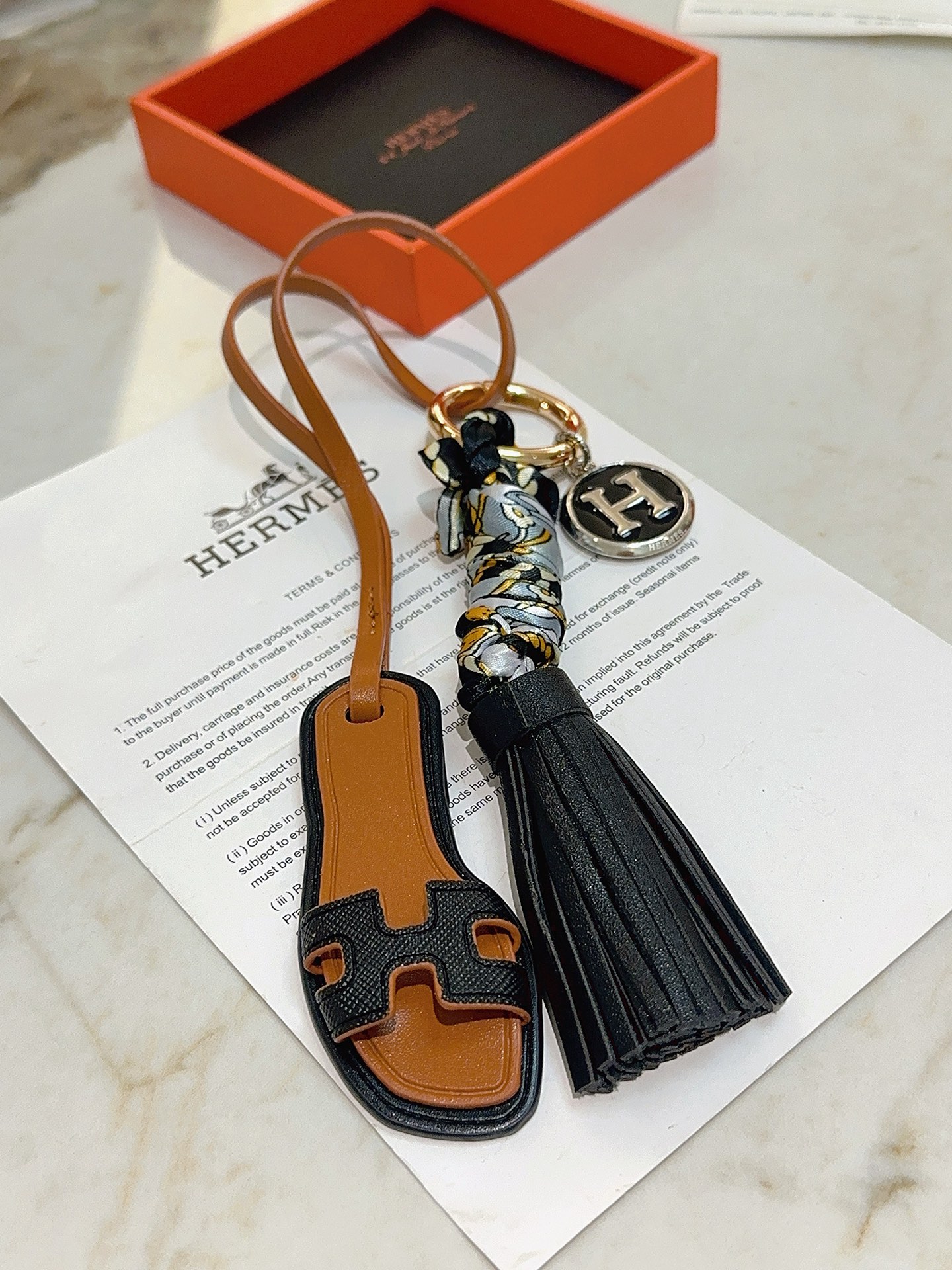 Hermes Oran Sandal Tassel H Logo Keychain Charm pingente metal dourado couro preto couro bronzeado metal prateado com esmalte preto seda cinza com estampa floral dourada e preta 12 cm de comprimento total