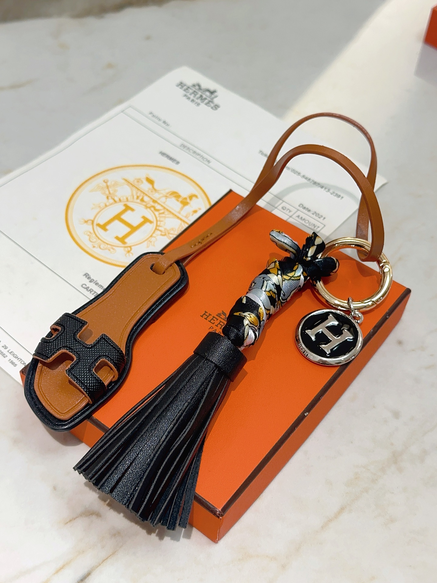 Hermes Oran Sandal Tassel H Logo Keychain Charm pingente metal dourado couro preto couro bronzeado metal prateado com esmalte preto seda cinza com estampa floral dourada e preta 12 cm de comprimento total