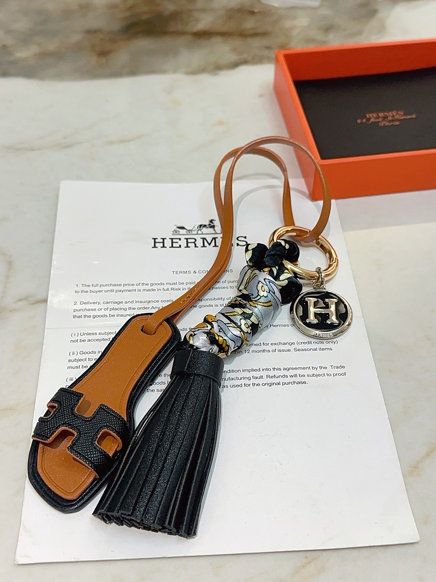 Hermes Oran Sandal Tassel H Logo Keychain Charm pingente metal dourado couro preto couro bronzeado metal prateado com esmalte preto seda cinza com estampa floral dourada e preta 12 cm de comprimento total