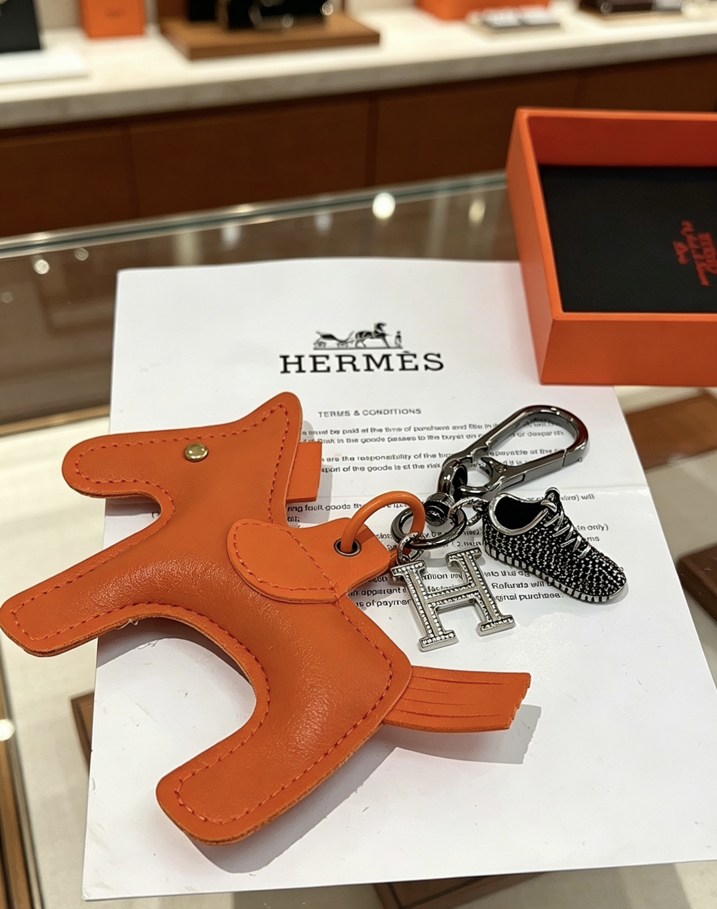 Hermes Rodeo H Logo Sneaker Charm Llavero colgante Herrajes de cuero naranja detalles en cristal transparente Charm de zapatilla en negro y plata 12 cm de longitud total