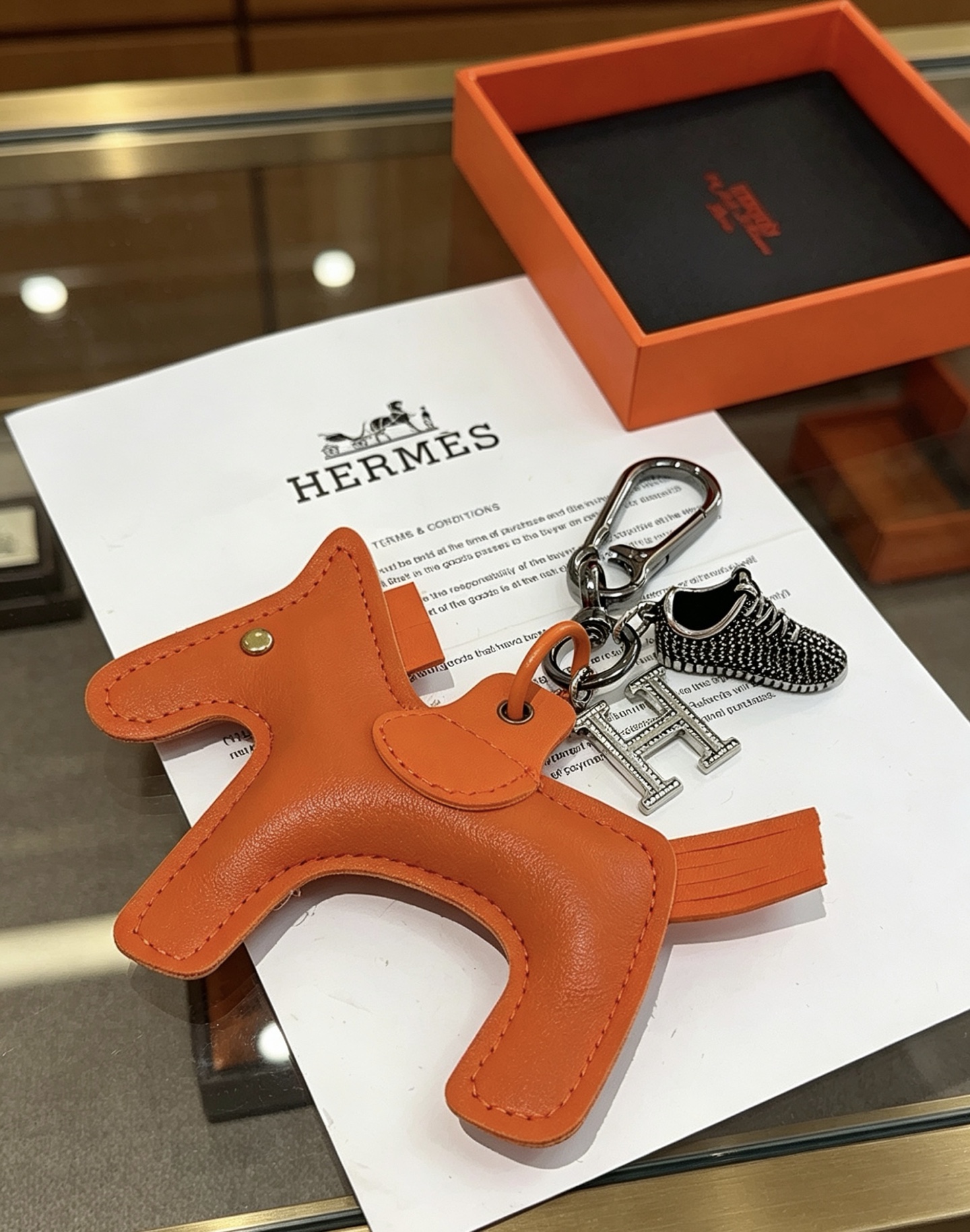 Hermes Rodeo H Logo Sneaker Charm Llavero colgante Herrajes de cuero naranja detalles en cristal transparente Charm de zapatilla en negro y plata 12 cm de longitud total