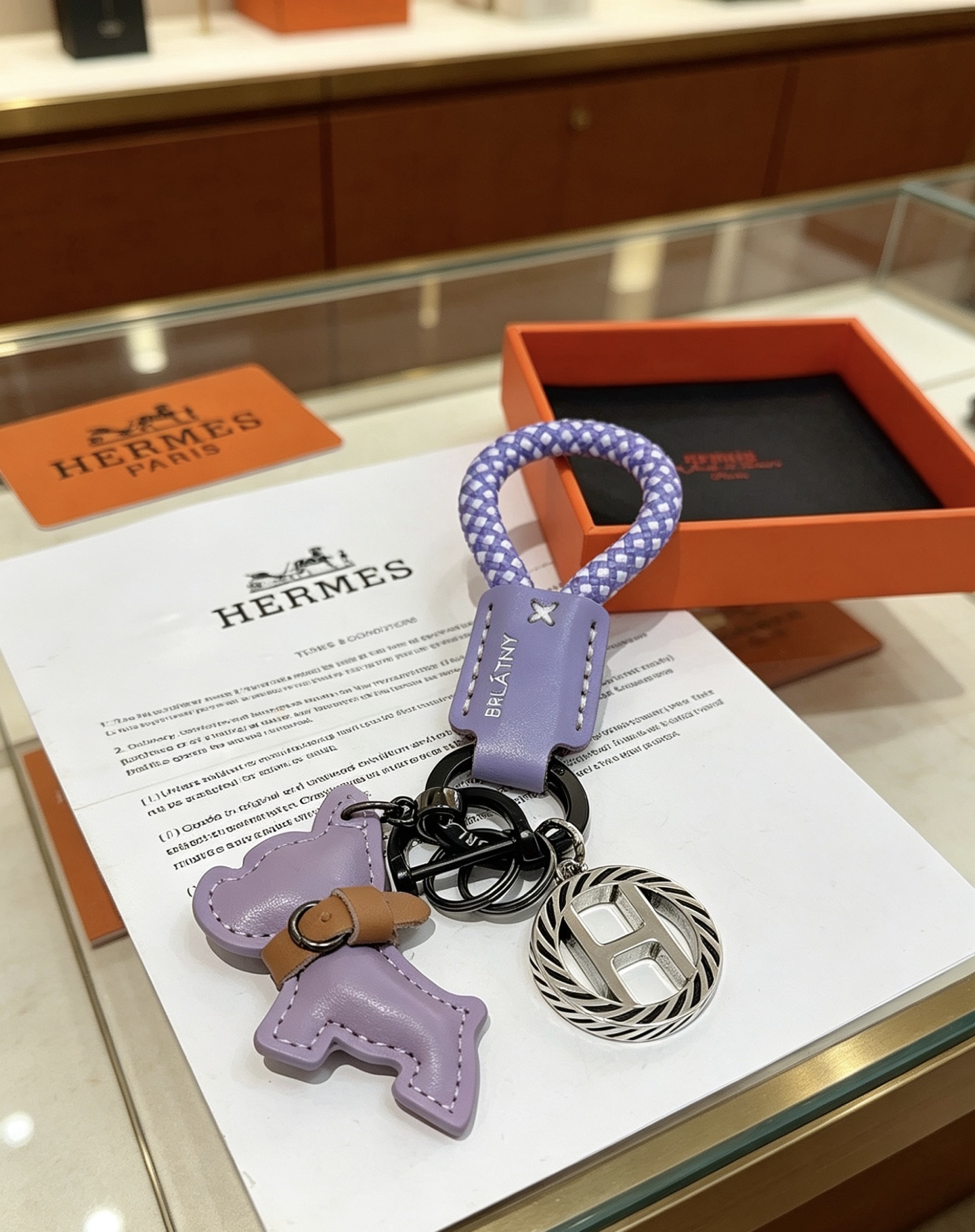 Hermes Rodeo H Logo Key Charm colgante Cuero morado lila Cordón trenzado morado y blanco Detalle de cuero bronceado Metal en tono plateado con detalles de esmalte negro Longitud total de 10 cm