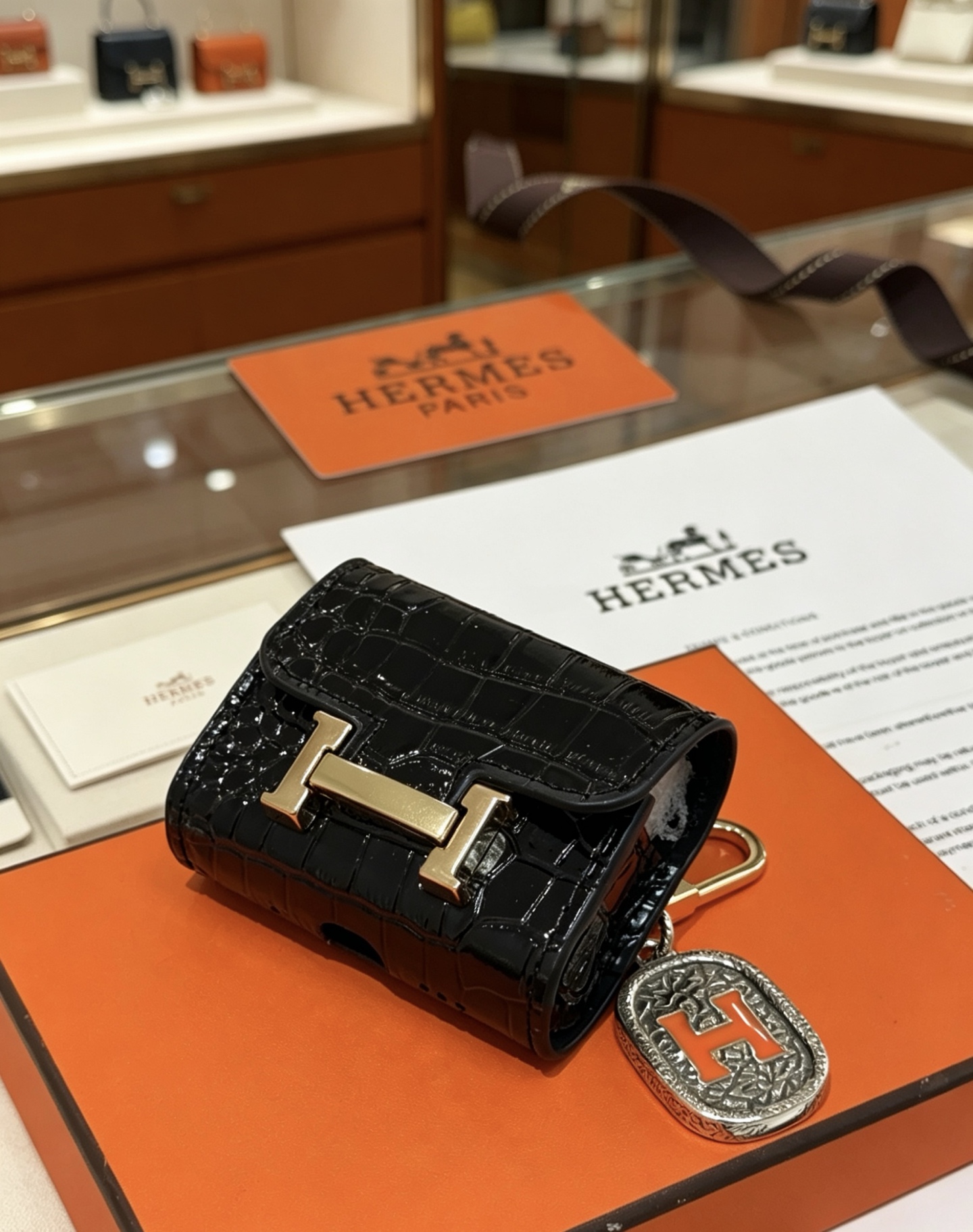 Hermes Constance Custodia per AirPods Borsa Ciondolo con ciondolo Pelle verniciata nera goffrata in coccodrillo Hardware in metallo color oro Ciondolo in metallo color argento con dettaglio H in smalto arancione Taglia unica adatta al ciondolo AirPods standard, lunghezza totale circa 8 cm